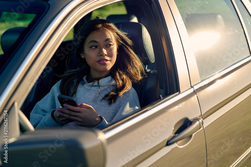 Young asian woman driving at golden hour, holding a smartphone and gazing out the window, reflecting on travel and connection during a peaceful road trip at sunset