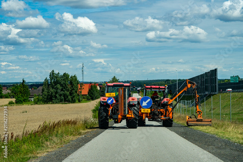 Obraz na plátne Two orange tractors with boom mowers attached driving on a paved service road next to soundproof barriers mowing grass along a highway under a cloudy blue sky