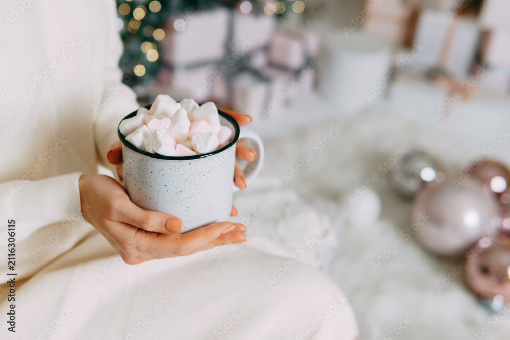 Obraz premium Woman holding cup of cocoa with marshmallow by Christmas tree. Cozy home atmosphere during winter holiday. Warm seasonal beverage for celebration and relaxation. Festive mood winter interior.