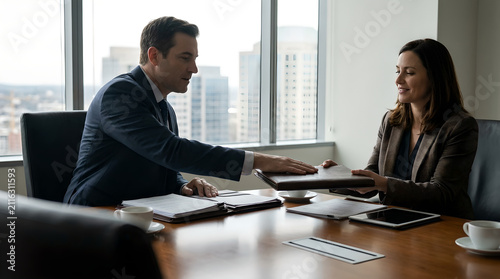 Business professionals sit across from each other at a polished conference table while exchanging a leather portfolio in a bright, modern corporate office.