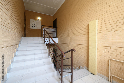 staircase in a residential building features white tiled steps, yellow brick walls, a dark metal handrail, an electrical panel, wall-mounted light fixture