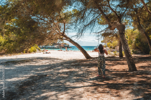 Fotografie View of a woman standing under pine trees on a sandy beach looking towards the turquoise sea in Ciutadella, Balearic Islands, Spain