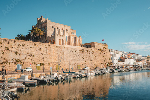 Fotografie View of the historic fortifications and buildings overlooking the harbor with moored boats in the calm water under a clear blue sky in Ciutadella, Balearic Islands, Spain