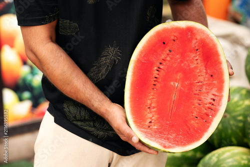 Man holding large halved ripe watermelon wrapped in plastic foil at local farmers market.