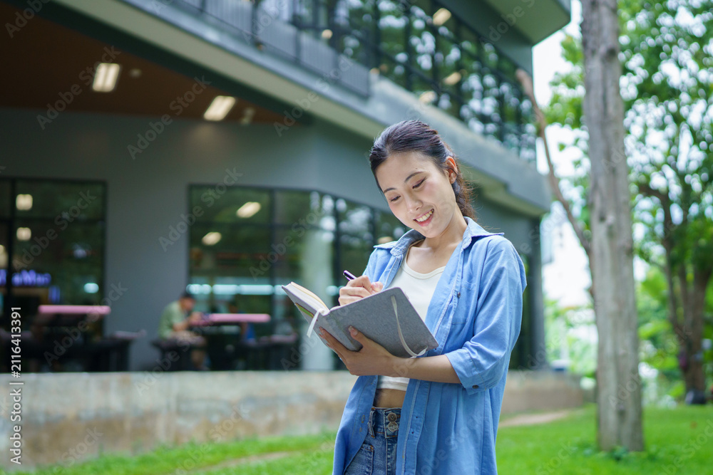 Fototapeta premium Education, university and woman in campus, Young asian woman smiles widely while tacking note. She standing in campus with her ready. Enjoying education outdoors with textbook