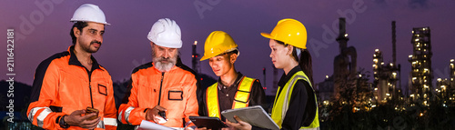 Industrial workers in safety gear review plans at a refinery site at dusk  wearing helmets and high-visibility clothing with plant lights glowing in the background.