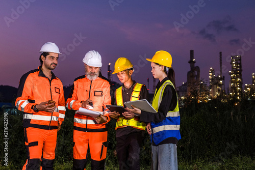 Industrial workers in safety gear review plans at a refinery site at dusk  wearing helmets and high-visibility clothing with plant lights glowing in the background.