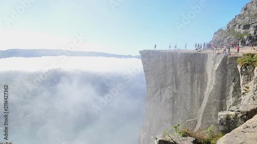 Norway - 28.07.2026: Hikers stand on Preikestolen plateau as morning fog fills the fjord below under clear sky.