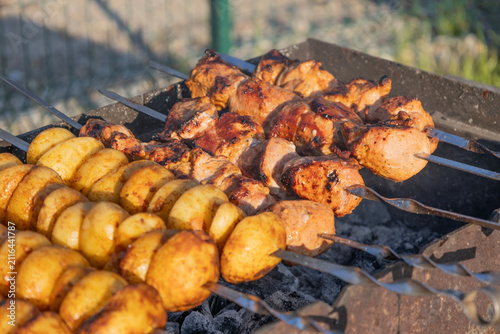 Delicious meat shashlik skewers and golden potatoes roasting on metal skewers over hot charcoal in a grill outdoors. Summer barbecue party food cooking process close-up photography.