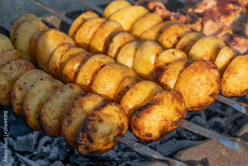 Delicious meat shashlik skewers and golden potatoes roasting on metal skewers over hot charcoal in a grill outdoors. Summer barbecue party food cooking process close-up photography.