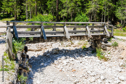Rustic wooden footbridge over a dry rocky creek in a lush evergreen forest, peaceful summer nature landscape with hiking trail and tranquil outdoor scenery.