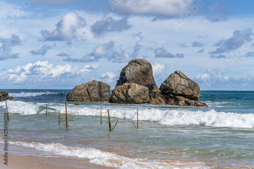 View of traditional stilt fishing poles in the turquoise waters of Ahangama Beach with large rock formations in the background in Galle, Southern Province, Sri Lanka.