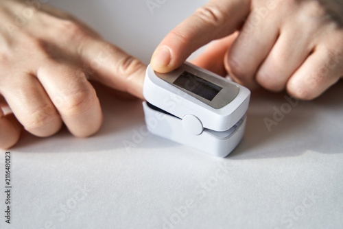 Obraz na plátne Pulse Oximeter Between Fingers Measuring Oxygen Saturation And Pulse Rate On White Table Top Close Up Portable Medical Device With Hands Adjusting Sensor For Home Monitoring