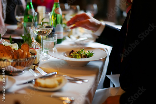 Guests are seated around a table enjoying a variety of food. A hand reaches for a dish that includes salad. Drinks are placed on the table. Soft lighting adds to the atmosphere.
