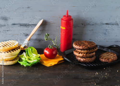 beef patties are grilled on a flat pan with fresh lettuce, cheese, tomato, and sauces nearby. The scene shows a rustic wooden table with various ingredients burger toppings ready to use.
