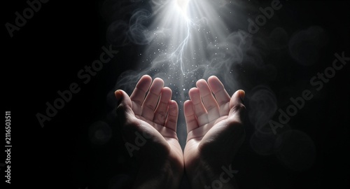Two human hands opened in prayer, receiving a dramatic divine light with smoky etherial effects and glowing dust particles, isolated against a dark black background to imply faith.