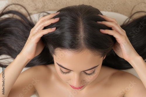 Top view of a young woman lying down, using hands to part her thick dark hair and show a clean healthy scalp line for spa wellness, beauty, and premium hair care concepts.