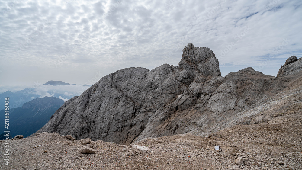 Fototapeta premium Panoramic aerial view of a lush alpine valley in the Italian Dolomites with towering limestone cliffs, dense forests, winding mountain roads, and dramatic storm clouds under a vibrant blue sky.