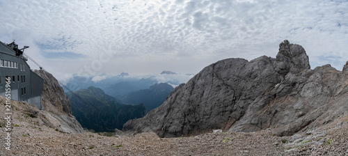 Panoramic aerial view of a lush alpine valley in the Italian Dolomites with towering limestone cliffs, dense forests, winding mountain roads, and dramatic storm clouds under a vibrant blue sky.