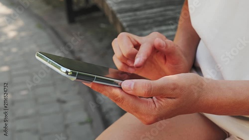 A person is sitting on a wooden bench in a park, looking at a smartphone. The sun is shining, and it is a warm afternoon