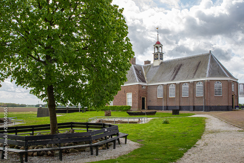 Schokland, Netherlands - 17 May 2015: View of the historic Enkhuizerkerk church on the former island of Schokland with a large green tree and circular bench under a cloudy sky.