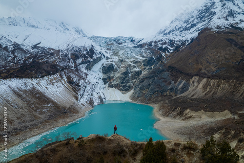 Aerial view of a person standing on a rocky ridge overlooking a turquoise glacial lake surrounded by snow-capped Himalayan mountains in Nepal.