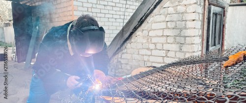 Welder wearing mask, goggles and hardhat sparks and smoke while fabricating steel on a construction site workshop, showcasing industrial safety gear, skill and heavy-industry metalwork.