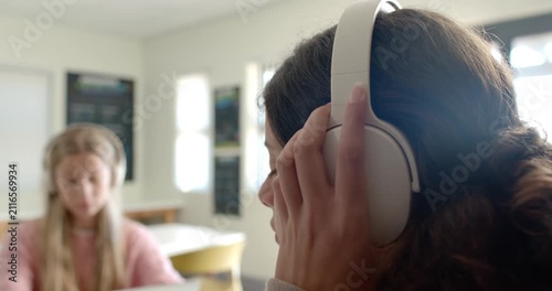 Two teen girls getting over-ear headphones placed by hand in classroom, adjusting fit studying