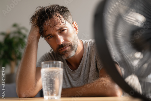 Exhausted person resting with their head on a table during a hot summer day, with a small fan blowing cool air nearby