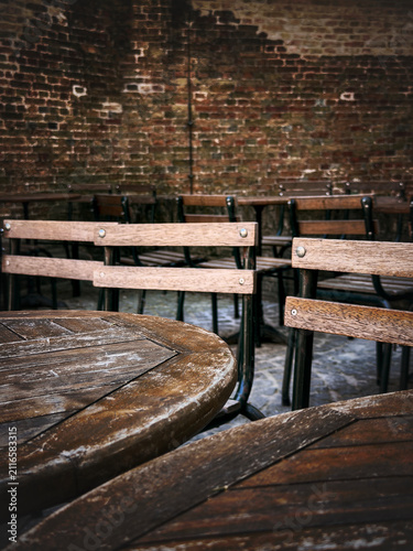 Close up of weathered wooden outdoor cafe tables and chairs arranged neatly against an old textured red brick wall in a European courtyard.