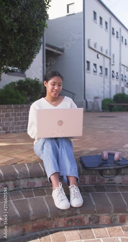 Vertical video: Asian child sitting on ledge at campus studying with laptop open, pack and headset