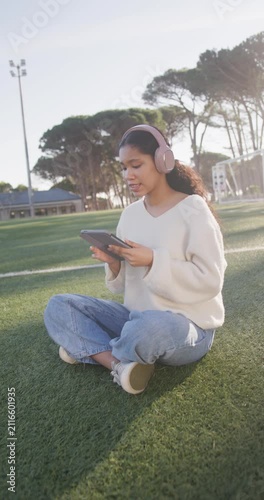 Vertical video: Girl child sitting on turf in pink headphones, tapping tablet and following audio