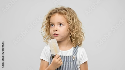 A delightful portrait of a young child with charming curly hair, looking directly at the camera with wide, expressive eyes, as they enjoy a creamy white popsicle. The child's innocent expression