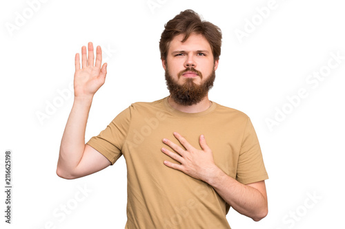 Fotografie Bearded young man with one hand on his heart and the other raised, making a solemn oath gesture, isolated on white background, concept of honesty, promise and sincerity