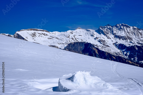 A stunning, expansive winter landscape showcasing a vast, untouched snowfield in the foreground, textured with subtle wind ripples and fresh ski tracks winding off into the distance. In the background