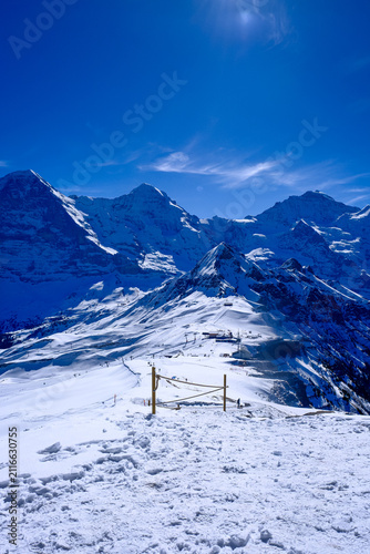 A breathtaking, wide-angle winter landscape capturing the iconic snow-covered peaks of the Eiger, Mönch, and Jungfrau mountains in the Bernese Oberland region of the Swiss Alps. Shot on a clear, sunny