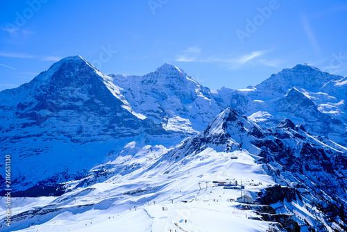 A breathtaking, wide-angle winter landscape capturing the iconic snow-covered peaks of the Eiger, Mönch, and Jungfrau mountains in the Bernese Oberland region of the Swiss Alps. Shot on a clear, sunny