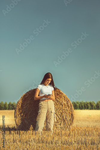 Fotografie A vintage portrait of a teenage girl finds a quiet moment in a nostalgic hayfield, absorbed in her smartphone