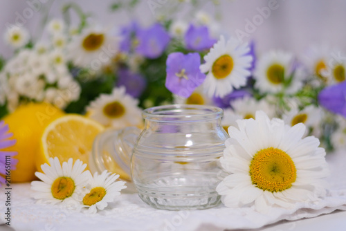 A clean empty glass jar stands among blooming daisies and delicate blue flowers, creating a fresh minimalist scene. Perfect for showcasing natural products, homemade preserves or honey.