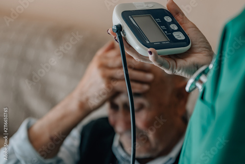 nurse holding digital blood pressure monitor while elderly patient looks worried, home medical care concept, hypertension check, healthcare visit