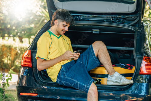 smiling young man in a summer outfit is sitting in the open trunk of a car, using his smartphone while traveling with a yellow suitcase on a sunny summer day