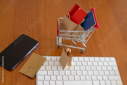 Electronic commerce items on a wooden desk. Including a miniature shopping cart filled with credit cards and paper bags. A smartphone. A keyboard. And an additional credit card