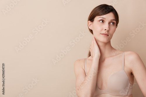 Foto A thoughtful woman stands in a soft beige studio, touching her neck with a calm expression