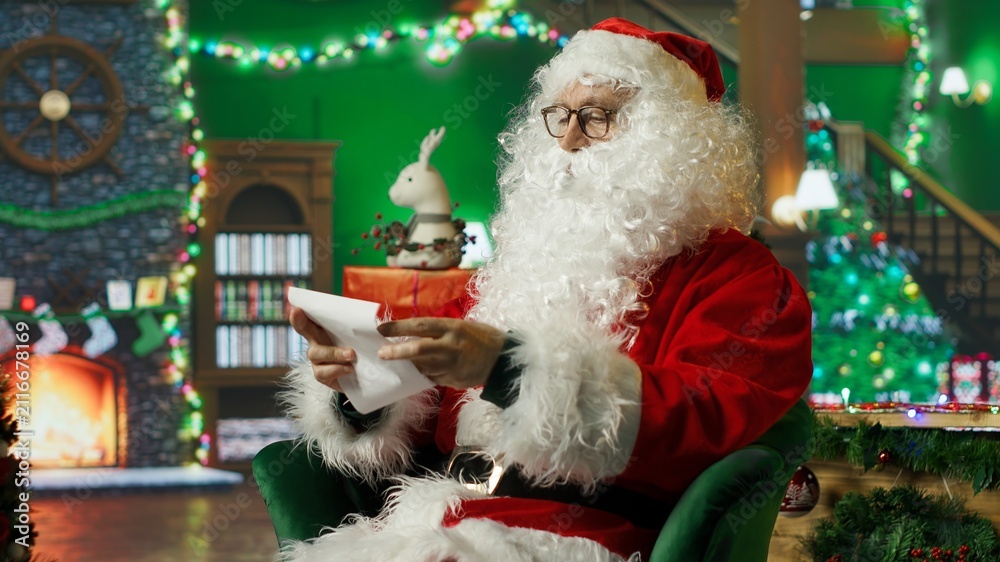 Obraz premium Senior man at home dressed as Santa Claus opens a letter from children wishing for gifts while wearing a red costume, then unpacks scrolls containing Christmas wish lists.