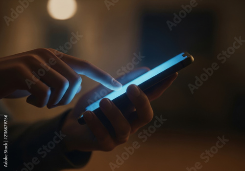 Human hand using modern smartphone in dark dimly lit room with glowing blue light reflecting on touch screen surface during nighttime use.