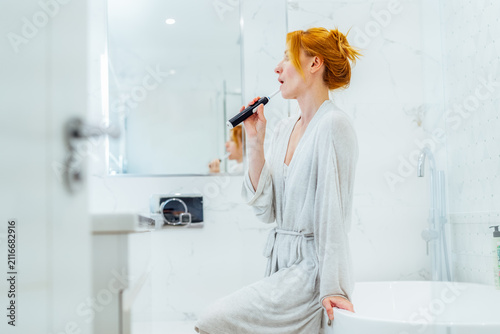 Middle aged woman cleaning her teeth with electronic toothbrush in her white modern bathroom. Using Sonic toothbrush for routine oral care. Modern home health care technology concept.