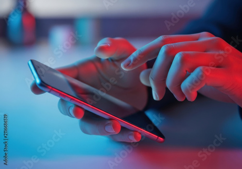 Person holding sleek smartphone while scrolling digital screen with finger in dimly lit environment featuring vibrant blue and red neon lighting glow.