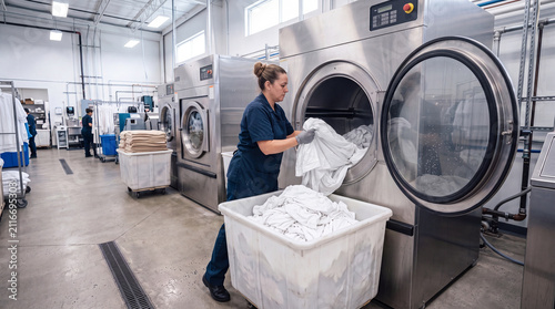 Housekeeper using large industrial washer in commercial laundry facility, basket filled with clean white sheets, bright workspace, modern professional washing equipment, cleanlines