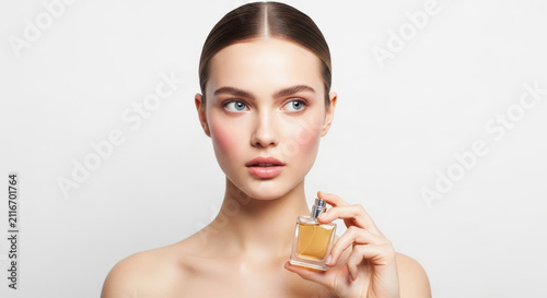 Elegant young woman with clean bare shoulders holding a perfume bottle looking directly at the camera with a neutral expression.