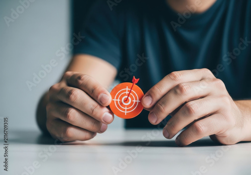 Hands holding small orange circle with target graphic and red arrow pointing at bullseye representing focused achievement precision and goal setting.